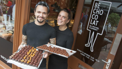 A man and a woman, each holding a tray of taster chocolates, stand in the doorway of Be Chocolat in Brighton