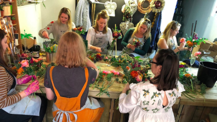 A group of women in overalls sit around a wooden table arranging flowers in a workshop at Between Two Thorns in Brighton