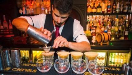 A bartender with a neatly groomed beard and wearing a white shirt with black vest and tie, is pouring a drink from a shaker into a cocktail glass through a strainer. Four filled cocktail glasses are lined up on the bar, with various liquor bottles in the background.
