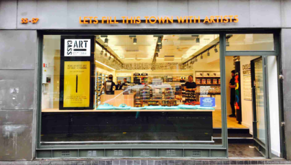 The exterior of Cass Art in Manchester; a cool grey building frames a warm interior filled with wooden floor and shelves of art supplies