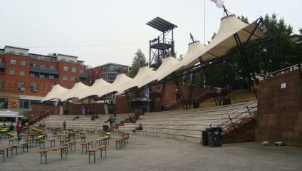 An outdoor public space with wooden benches and tables, adjacent to steps and covered by a series of large, white canopy structures supported by black metal frames. Surrounding the area are multi-story buildings and greenery.