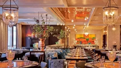 A luxurious restaurant interior featuring elegant chandeliers, a mix of contemporary and classic decor, and tables set with crystal glasses and fine china. Large floral arrangements and vibrant artwork add color to the warmly lit space.