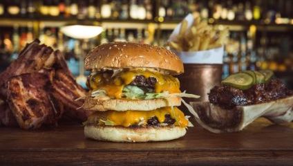 A double cheeseburger with lettuce and melted cheese on a sesame seed bun is centered in the image. To the left, there are slices of grilled meat. To the right, there are French fries in a copper container and a dish with pickles and ribs. A bar setup is visible in the background.