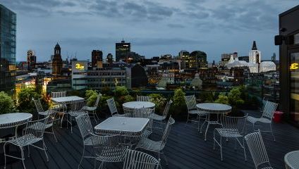 A rooftop terrace with white metal tables and chairs overlooks a cityscape at dusk. The skyline features various buildings and skyscrapers with some lights turned on, under a cloudy sky. The terrace has greenery along the railing and a glass-walled structure to the right.