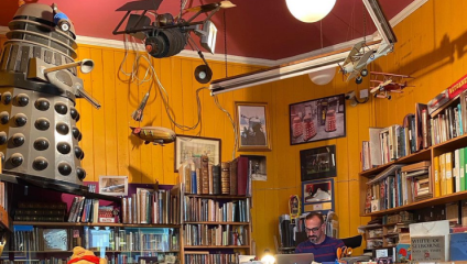A man is working at a cluttered desk in a room filled with bookshelves. The shelves are lined with various books and framed pictures. Hanging from the ceiling are several model airplanes, and a large Dalek model stands prominently in the corner.