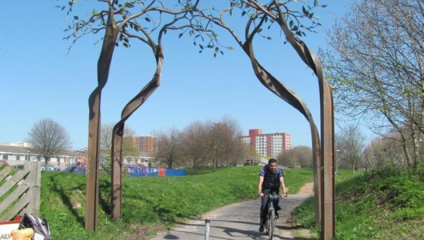A cyclist rides under an artistic metal arch designed to resemble tree branches on a sunny day. The path is surrounded by grassy areas and trees with a clear, blue sky above. Buildings and a fenced area are visible in the background.