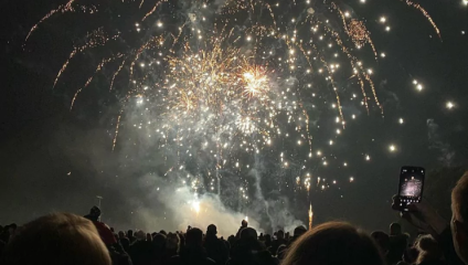 Yellow and orange fireworks light up the sky above thousands of onlookers below, some filming with their phones and some lighting flares at the Donwnend Roundtable Fireworks display in Bristol in 2023