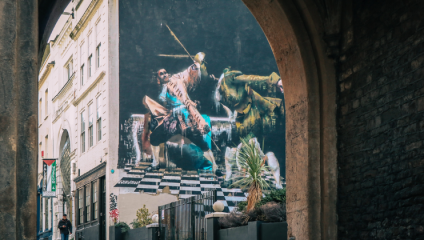 A vibrant and detailed mural is visible beneath a stone archway, depicting a knight on horseback with a vibrant mix of colors, creating a dramatic effect. The mural is painted on the side of a building, surrounded by urban scenery and greenery.