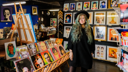 A woman with blue hair, wearing a black dress and hat, stands looking at the camera surrounded by art prints in the Egoiste Gallery in Manchester’s Afflecks Palace