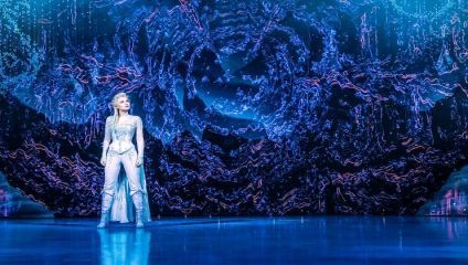 A performer dressed in a sparkly, light blue costume stands confidently on a stage. The background is a dazzling, abstract display of blue and purple lights, creating an immersive, magical atmosphere that seems to swirl around the performer.