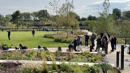 A park scene with people strolling on pathways, sitting on benches, and socializing. There is abundant green vegetation, flowers, and scattered trees. A group is gathered near a grassy area while someone is seen taking photos. The sky is partly cloudy.