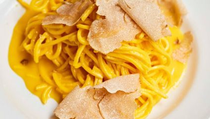 A close-up image of a plate of spaghetti with a creamy yellow sauce, topped with thinly sliced truffle shavings. The dish is presented on a white plate, showcasing the rich texture of the pasta and the luxurious truffle topping.
