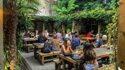 People are seated at large wooden picnic tables in an outdoor garden area. The setting is lush with greenery, palm trees, and shrubbery. Some individuals are drinking and talking, while others are using laptops. The space has a relaxed and social atmosphere.