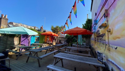 A vibrant outdoor seating area filled with wooden picnic tables and benches. Colorful umbrellas provide shade, and rainbow flags are strung overhead. A colorful mural decorates the adjacent wall, adding to the lively atmosphere. It's a sunny day with a clear blue sky.