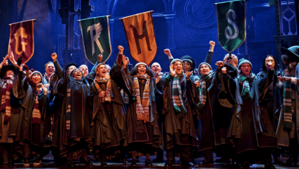 A group of people dressed in Harry Potter-themed robes and scarves cheer enthusiastically on stage. They hold banners with letters 'G', 'R', 'H', and 'S'. The background features a dimly lit, elaborate set with gothic-style arches.