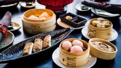 A variety of dim sum dishes are presented on a table. The image includes bamboo steamers with dumplings, shumai, and other assorted items. Plates and bowls containing dipping sauces accompany the food. The table setting includes utensils and cloth napkins.