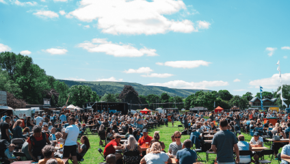 A large crowd of people enjoying a sunny day at an outdoor festival. They are sitting at picnic tables spread across a grassy area, with food stalls and other festival structures in the background. The scene is set against a backdrop of trees and distant hills.