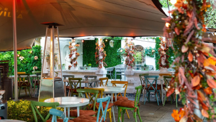 An outdoor cafe with colorful chairs and round tables under a canopy. Autumn-themed decorations hang around the space, and a patio heater stands nearby. The backdrop features lush greenery and string lights, creating a cozy and inviting atmosphere.