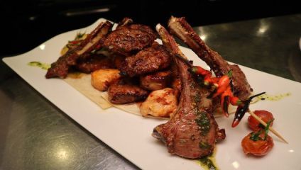 A white rectangular plate with a variety of grilled meats, including lamb chops, chicken pieces, and skewered vegetables, garnished with herbs and served with a side of cherry tomatoes.