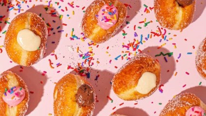 A pink surface is covered with sugar-coated donuts filled with cream, pink icing, and chocolate filling. Colorful sprinkles are scattered around and on top of the donuts, creating a festive, playful appearance.