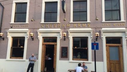 Street view of Chesham Arms pub with people seated and standing outside. The building is light purple with white trim, featuring large windows and wooden doors. Signs for Local Ales and Fine Wines flank the pub name. A blue no entry sign is visible.