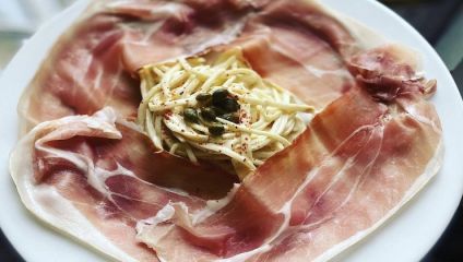 A plate with thin slices of prosciutto surrounding a central portion of pasta topped with capers and a sprinkle of red seasoning. The spaghetti is lightly coated in a creamy sauce and attractively arranged.