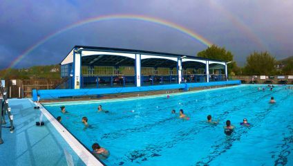 An outdoor swimming pool with multiple swimmers is shown under a partly cloudy sky. A double rainbow stretches across the sky, arching over the pool. In the background, there is a covered seating area and green trees. The scene is vibrant and lively.