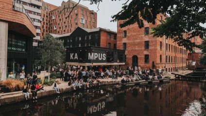 A lively urban scene by a canal with people socializing. Modern and historic buildings form the backdrop, including a black building with large white letters spelling KAMPUS. Trees frame the view, and reflections of the buildings are visible in the canal.