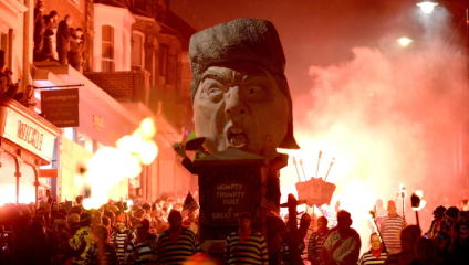 During the Lewes Bonfire Night celebrations, dozens of men dressed in black-and-white striped jumpers and red beanies lead of a huge effigy of President Donald Trump through the streets. The night sky is lit orange and red by the flames of torches carried by men in the procession, and hundreds of spectators watch from the pavement, with some even climbing into windows and onto roofs to see