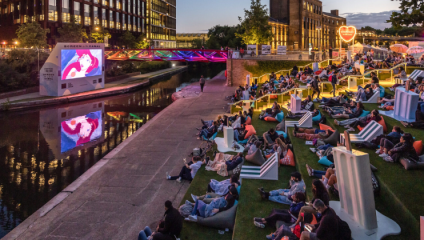 People are seated on grass, loungers, and bean bags by a canal watching an outdoor movie at dusk. A colorful bridge and illuminated signs add to the lively atmosphere. Buildings and trees are visible in the background.