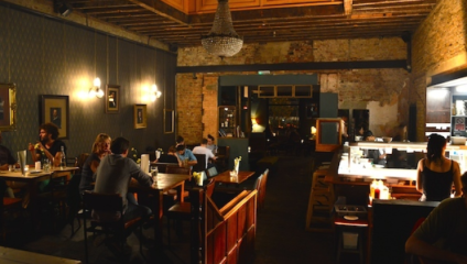 A cozy restaurant interior with exposed brick walls and dim lighting. Diners sit at wooden tables, engaged in conversation, while a chandelier hangs from the wooden ceiling. Lights provide a warm ambiance, and the overall atmosphere is intimate and inviting.