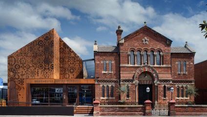 Image shows the Manchester Jewish Museum, featuring an older brick synagogue building beside a modern extension with ornate perforated metalwork. The old and new structures are seamlessly integrated, set against a partly cloudy blue sky.