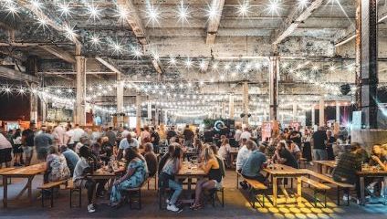 A bustling indoor food market, filled with people eating and socializing at communal tables. The space is decorated with numerous string lights hanging from the ceiling, creating a warm and lively atmosphere. Various food stalls are visible in the background.