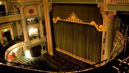 A luxurious, ornately decorated theater with a large stage featuring a closed, green curtain with gold trim. The interior includes grand balconies and intricate architectural details in green and gold. The theater is empty, with seats facing the stage.