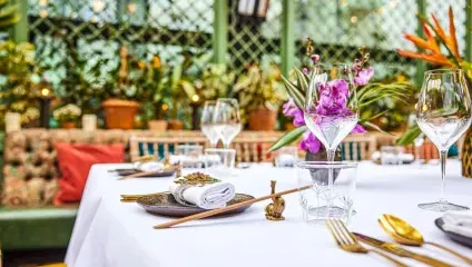 A beautifully set dining table in a vibrant garden-themed restaurant with a white tablecloth, elegant wine glasses, gold cutlery, and floral centerpieces. Lush green plants, colorful flowers, and decorative pillows adorn the background.