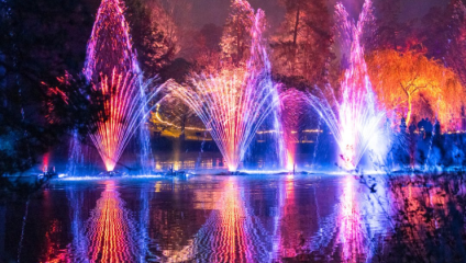 Three multi-pronged water jets fly up from the lake at Mannings Heath Golf Club, the sky illuminated red, blue and orange by the fireworks above. The beautiful scene is reflected in the water of the lake.