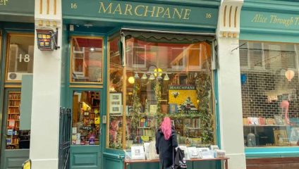 A person with pink hair and a black coat stands outside Marchpane, a bookstore with a green façade. The display window is filled with books and various decorations. A table with books for sale is set up in front of the shop.