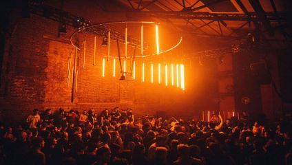 A large crowd of people is dancing and socializing in a dimly lit nightclub. Above them, red fluorescent tube lights hang from the ceiling, casting a warm, vibrant glow. Exposed brick walls and industrial beams add to the club's aesthetic.