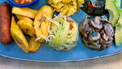 A rectangular platter holds a colorful assortment of foods, including beans in a blue bowl, triangular potato pieces, greens, avocado slices, mushrooms, fried plantains, and a vegetable medley. Below are three smaller dishes featuring fish, fried plantains, and fried chicken.