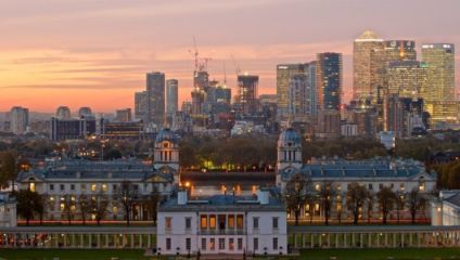 View from Greenwich Park at dusk, highlighting the historic Queen's House in the foreground, with two domes of the Old Royal Naval College flanking it. The modern Canary Wharf skyscrapers form the backdrop, standing against a colorful sunset sky.