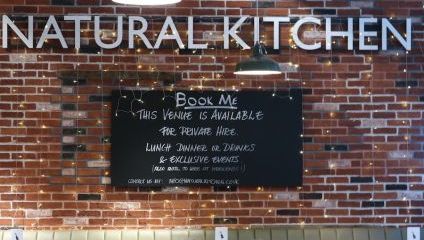 Restaurant interior with a sign above reading NATURAL KITCHEN. Below is a blackboard with text: BOOK ME. This venue is available for private hire. Lunch, dinner or drinks & exclusive events. (Also available to hire as B.Y.O restaurant or bar)..