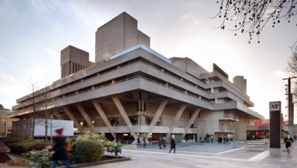 The image shows the exterior of the National Theatre in London, a Brutalist-style building with angular concrete structures and overhanging terraces. People are walking around the plaza in front, and trees with bare branches are visible under a cloudy sky.