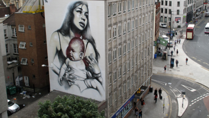 A large mural on the side of a building depicts a woman holding a baby. The painting is monochromatic and covers several stories of the building. The scene is set in an urban area with pedestrians, buildings, and a bus visible on the street below.
