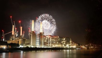 A night view of a cityscape, featuring an industrial building with several tall smokestacks. Fireworks are exploding above the building, illuminating the sky. Cranes and modern buildings are also visible along the waterfront, reflecting on the water.