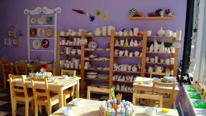 The interior or the Painting Pottery Cafe in Brighton; pottery materials, paints and pencils are laid out on small wooden tables, and pottery pieces are arranged on a set of shelves