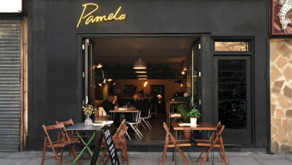 The doors of London bar Pamela are wide open, with table and chairs arranged both inside and outside. The black storefront is decorated with a thin yellow neon sign displaying the word Pamela in an italic font