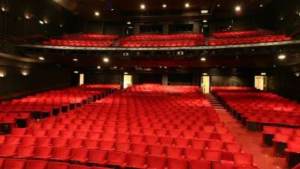 A large, empty theater with rows of red seats facing a stage. The interior is dimly lit, highlighting the seats which are organized neatly on the ground level and balcony. The walls are dark with acoustic panels, and small lights are scattered on the ceiling.