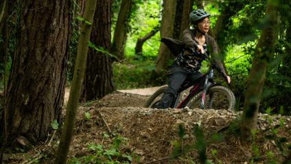 A person wearing a helmet, jacket, and gloves enjoys riding a bike on a dirt trail through a dense, green forest. The trail is surrounded by tall trees and lush foliage, and the person appears focused and engaged with the ride.