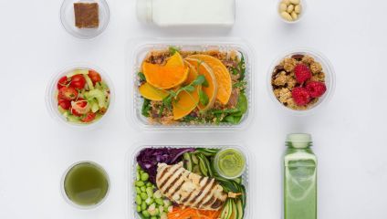 A flat lay image of a healthy meal set: two containers with salads (one topped with grilled chicken and the other with orange slices), two small cups of drinks, a white bottle, a green bottle, small bowls of cherry tomatoes, nuts, and raspberries with granola.