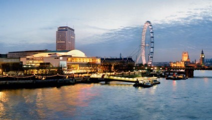 Evening view of the South Bank, London. The River Thames is in the foreground, with buildings lit up along the river. The London Eye is prominently featured and illuminated, while Big Ben and the Houses of Parliament are visible lit up in the background.